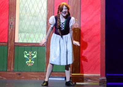 A student wearing a dress sings in front of a rustic indoor wall in the Priestnall School Frozen Jr Production.