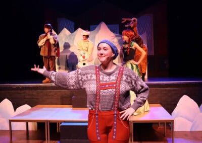 A student dressed as a lumberjack stands smugly with their arm extended, while Anna, Kristoff, Olaf and a reindeer stand in the background in the Priestnall School Frozen Jr Production.